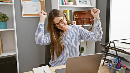 A cheerful woman in glasses celebrates success at her workplace with a laptop and stationery in a...