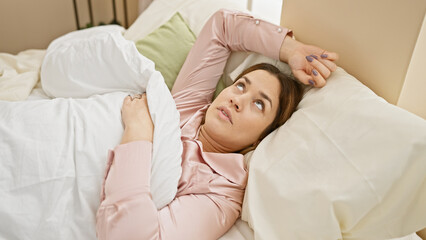 A pensive woman lies in bed gazing away, with her stylish pajamas, cozy bedroom setting, and thoughtful expression.