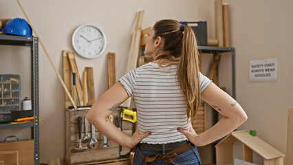 Confident young woman with ponytail stands in carpentry workshop, hands on hips, tools and wood...