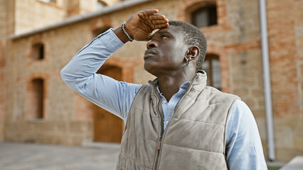 A contemplative african man standing outdoors in a city, shielding his eyes from the sun while...