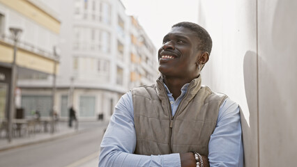 Smiling african man leaning against a wall on a sunny urban street wearing casual clothes and a vest