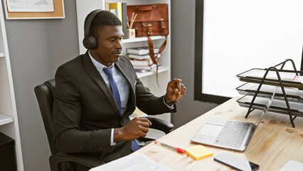 Handsome black man in a suit working indoors at his modern office desk with laptop and headphones