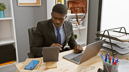 An african man in a suit works at his laptop in a modern office, phone and tablet also in use.