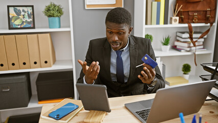 African businessman holding credit card looking perplexed in office.