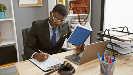 African american businessman in office analyzing document and taking notes with laptop and gavel...