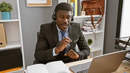 A professional african american man in a suit with headphones works on his laptop in a modern...