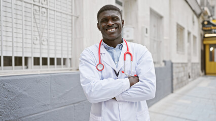 Smiling black man in white lab coat with stethoscope standing confidently outside urban clinic.