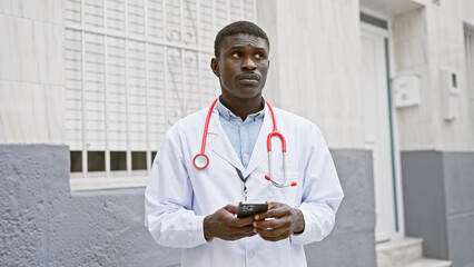 A professional african american man in a lab coat with a stethoscope stands outside a hospital...