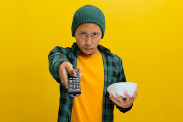 Annoyed Asian man, dressed in a beanie hat and casual shirt, switches TV channels using a remote...