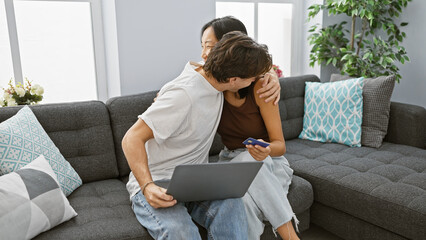 Interracial couple embracing in living room with laptop and credit card, signifying online shopping...