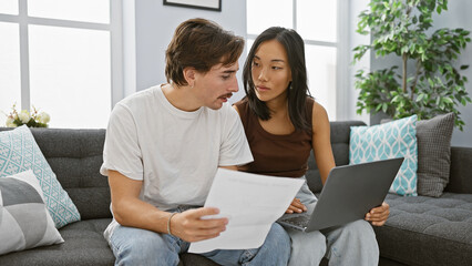 Interracial couple discussing paperwork in their modern living room, with a woman holding a laptop...
