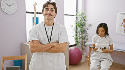 Fototapeta premium Confident male therapist smiling at camera with a female patient using dumbbells in a bright rehab center.