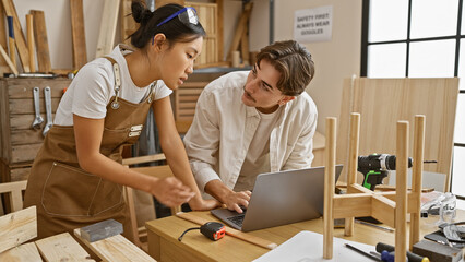 Two carpenters, a man and a woman, collaborate in a workshop, discussing work on a laptop...