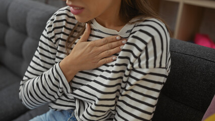 Young hispanic woman with hand on chest, wearing striped shirt, indoors at home, showing gesture of...