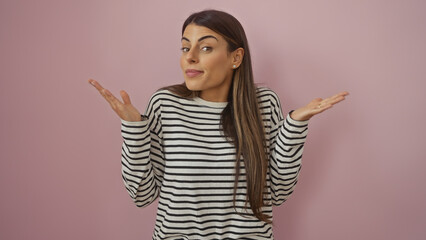 A young hispanic woman in a striped shirt expressing uncertainty against a pink wall.