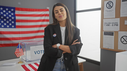 Confident young hispanic woman with arms crossed stands in an american polling center decorated...