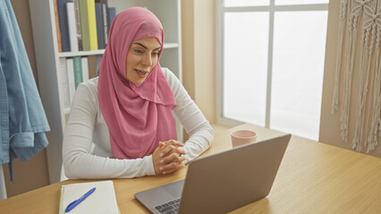A thoughtful woman in a hijab engages with a laptop in a bright home office setting.
