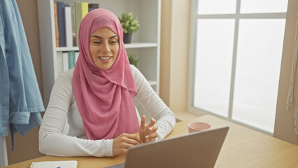Smiling woman wearing a pink hijab using a laptop at a bright, cozy home office.