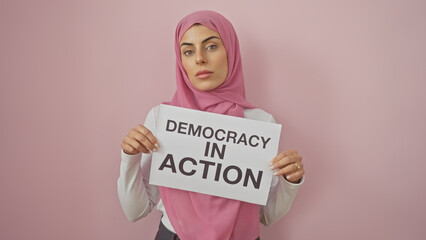 Confident muslim woman holding a 'democracy in action' sign against a pink background, representing...