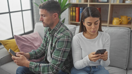 A man and woman sit apart on a sofa engrossed in their phones, highlighting modern indoor disconnection.