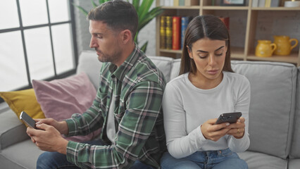 A man and woman sit apart on a sofa engrossed in their phones, highlighting modern indoor...