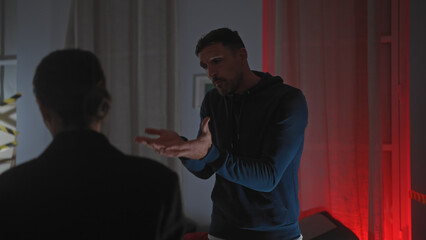 A man gestures while explaining himself to a policewoman in a dimly-lit indoor crime scene.