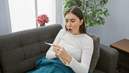 Shocked hispanic woman holding thermometer at home on couch