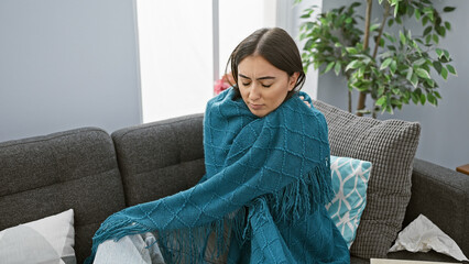 Hispanic woman feeling unwell wrapped in a blanket in a cozy home interior.