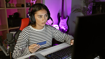 A focused hispanic woman holds a credit card while gaming in a neon-lit home room at night