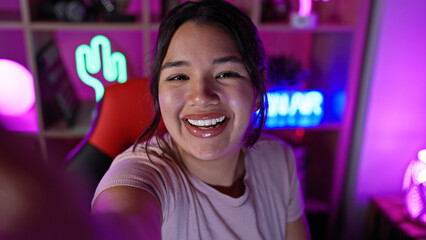 A happy young hispanic woman taking a selfie in a vibrant gaming room at night, with neon lights...