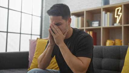 A distressed young hispanic man covers his face with hands, sitting in a modern apartment living...