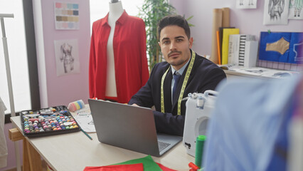 A handsome hispanic man with a beard works on a laptop in a colorful tailor shop surrounded by...