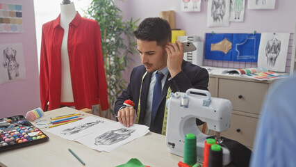 Handsome hispanic man listening to voicemail in a tailor shop with fabric sketches and a sewing...
