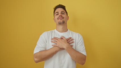 Handsome young hispanic man with a beard in a white t-shirt, hands on chest against a yellow...