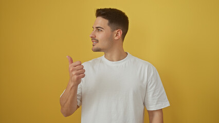 Handsome young hispanic man pointing with thumb to side against yellow isolated background, portrait