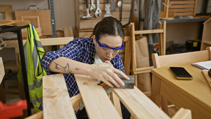 A focused woman sanding wood in a carpentry workshop surrounded by tools and furniture.
