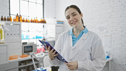 Smiling young caucasian woman in lab coat holding clipboard in modern laboratory setting