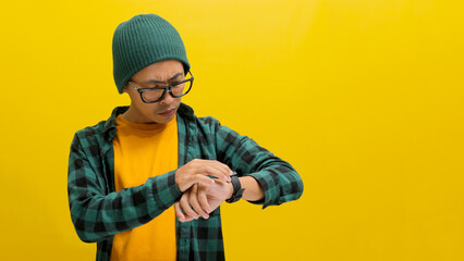 Asian man in a beanie and casual shirt checks the time on his wristwatch, isolated on a yellow...