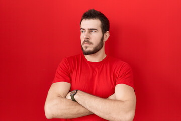Young hispanic man wearing casual red t shirt looking to the side with arms crossed convinced and confident