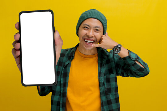 Asian man in a beanie hat and casual shirt shows a phone with a blank white screen to the camera while making a call-me gesture. Isolated on a yellow background