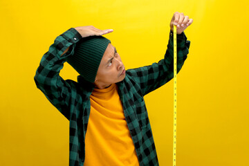 An Asian man, dressed in a beanie hat and casual clothes, measures his height using a yellow...