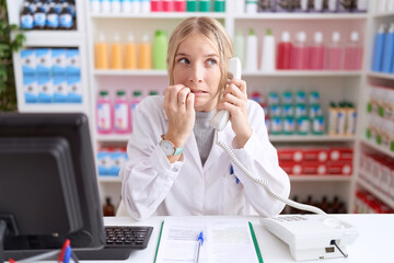 Young caucasian woman working at pharmacy drugstore speaking on the telephone looking stressed and nervous with hands on mouth biting nails. anxiety problem.
