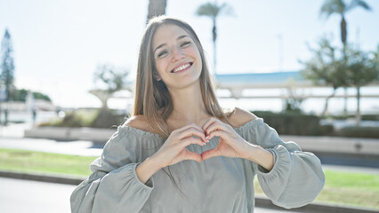 A young blonde woman forms a heart shape with her hands outdoors on a sunny city street.