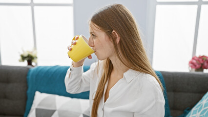 A beautiful woman drinking from a yellow mug, relaxed in a modern living room setting.