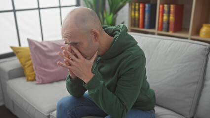 A concerned bald man with a beard sitting on a couch indoors looking stressed, covering his face with his hands.
