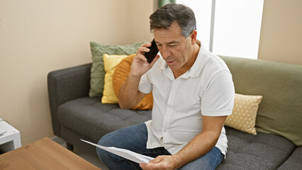 Mature man sitting on couch at home talking on phone holding paper concerned expression