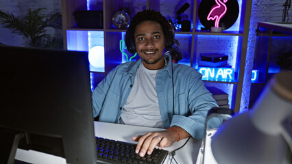 Handsome mixed race man with curly hair smiling in a dark gaming room at night, showcasing modern...