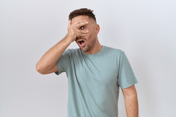 Hispanic man with beard standing over white background peeking in shock covering face and eyes with...