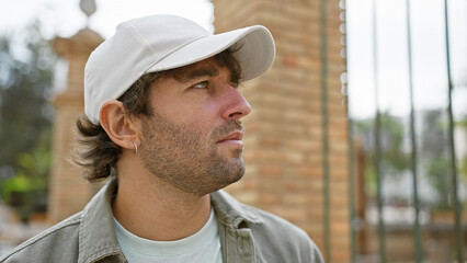 Side view of a contemplative young man with a beard wearing a white cap and casual clothing against...