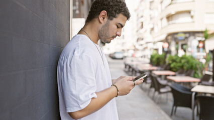 A young man with a beard using a smartphone on a sunny city street with outdoor cafÃ©s in the...